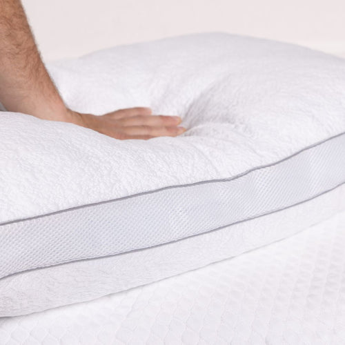 Close-up of a person's hand touching a white mattress topper with gray stitching on a light background.