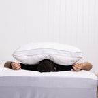 Person lying on a bed with a large white pillow over their head against a plain background