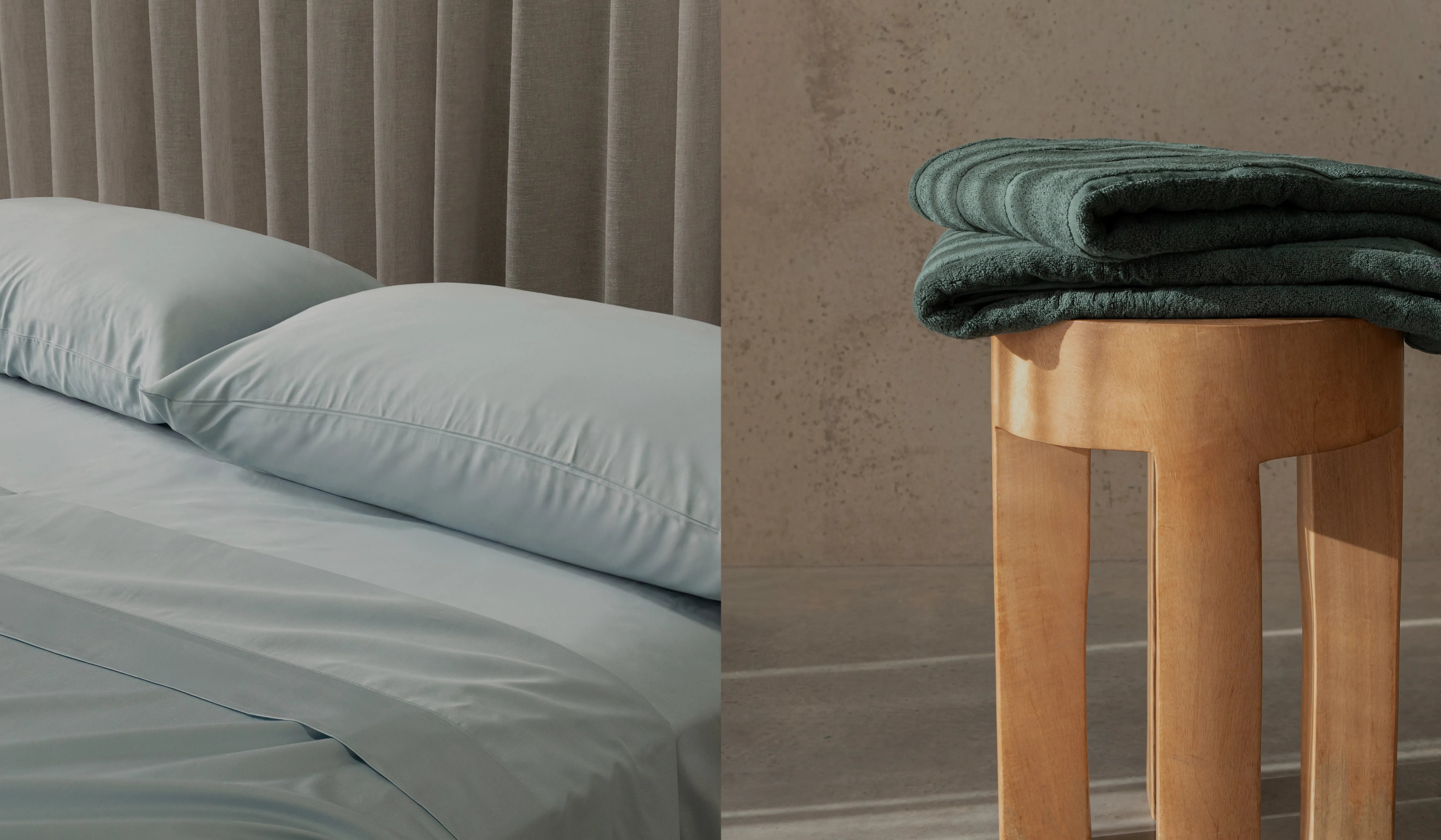 Bedding set with light blue bedding and pillows on a bed, and folded green blanket on a wooden stool against a neutral wall.