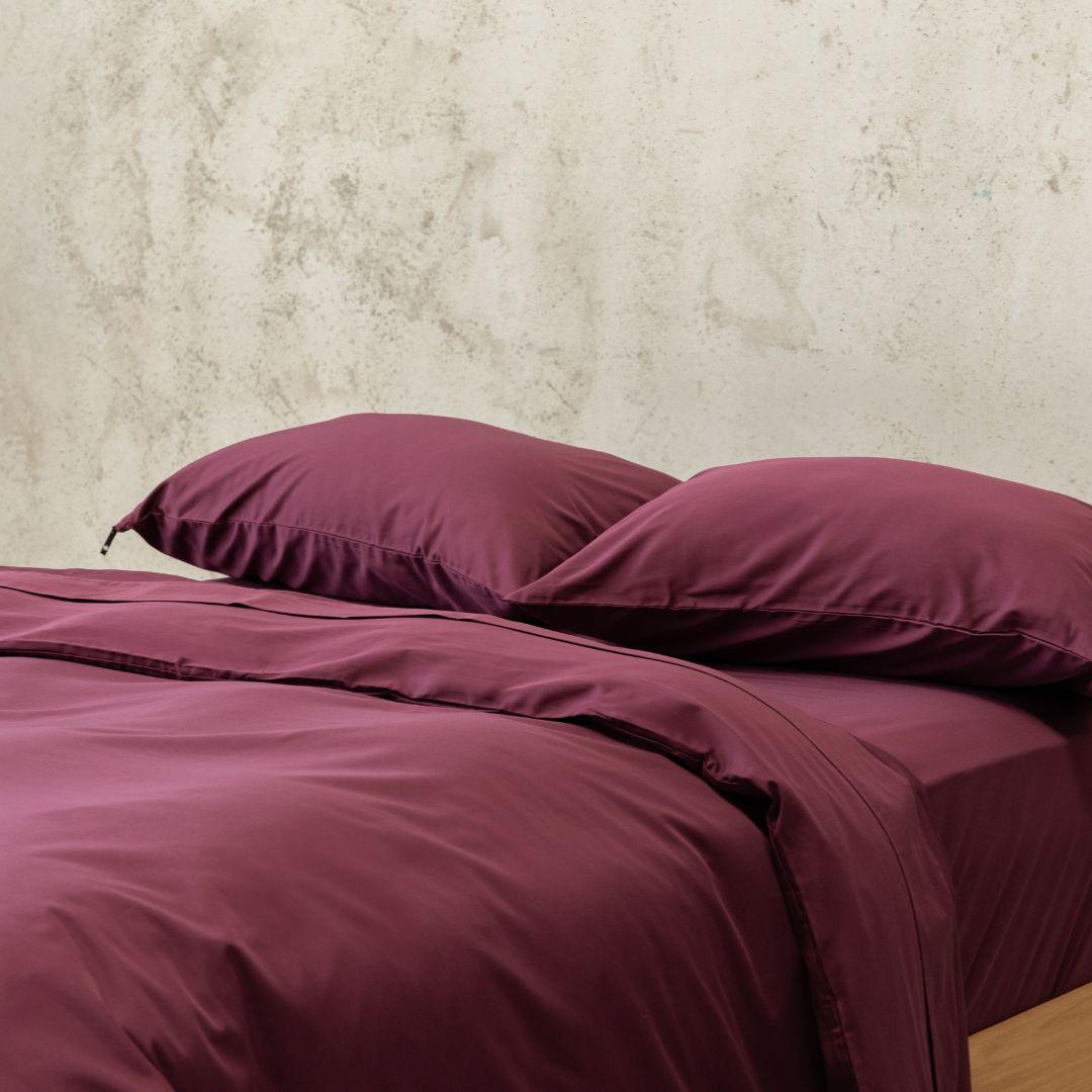 Burgundy bedding set with two pillows on a bed against a neutral wall.
