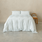 Light blue gingham quilt cover set with matching pillowcases displayed on a neatly made bed, styled in a minimalist bedroom with a woven wooden chair and neutral textured wall