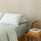 Close-up of a light blue gingham quilt cover and pillowcases layered with soft blue fitted sheets, styled beside a woven wooden chair in a modern bedroom setting