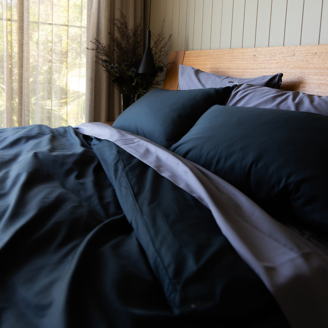 Bed with dark bedding and pillows in a room with natural light filtering through curtains.