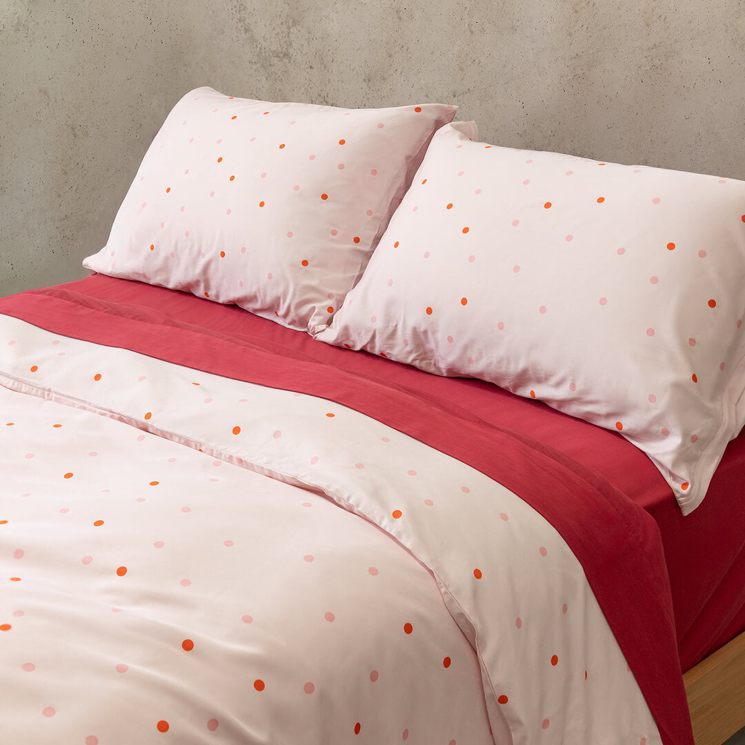 Pink and red polka dot quilt cover set on a bed with red sheets and a concrete wall background.