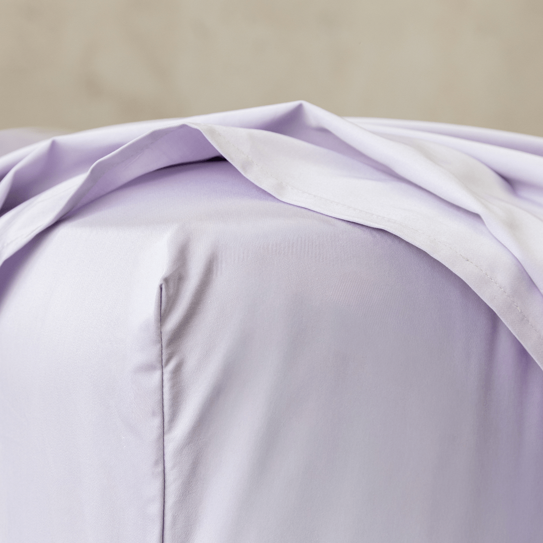 Close-up of a light lavender fitted sheet stretched over a mattress corner, showing the smooth fabric and stitched finish.