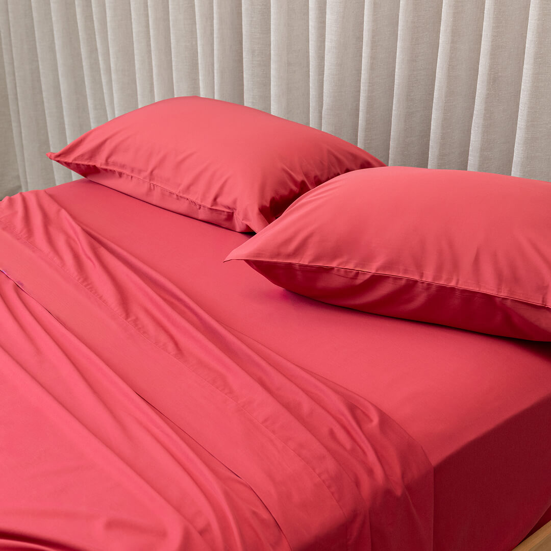Red bed sheets and pillows on a bed with light gray curtains in the background.