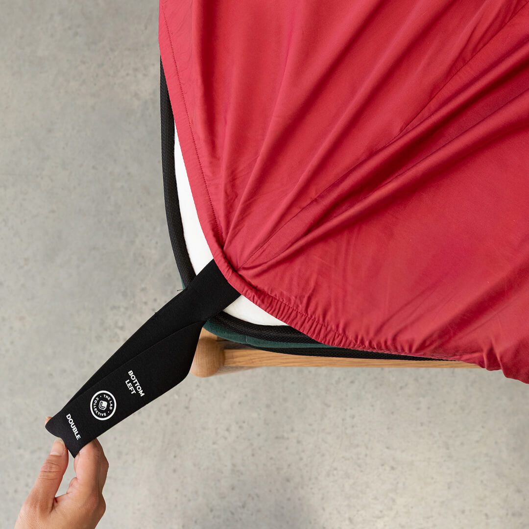 Close-up of a red fitted sheet and black labelled corner loop strap being pulled over the corner of a mattress with hand.