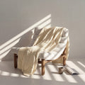 Beige cooling blanket draped over a wooden chair with a cup and book on a white surface.
