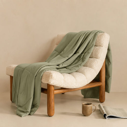 Sage green chair with a green blanket draped over it, accompanied by a mug and newspaper on a beige surface.