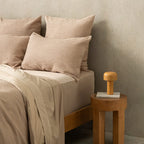 Beige bedding with pillows on a bed next to a wooden side table with a lamp.