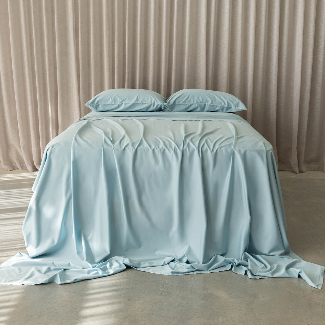 Light blue bedspread on a bed with matching pillows against a beige curtain background.