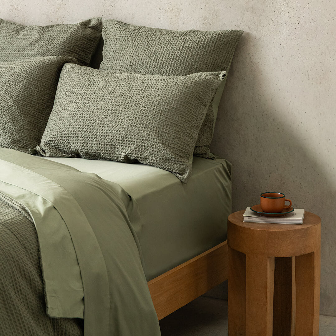Bed with green bedding and pillows next to a wooden side table with a cup and book.