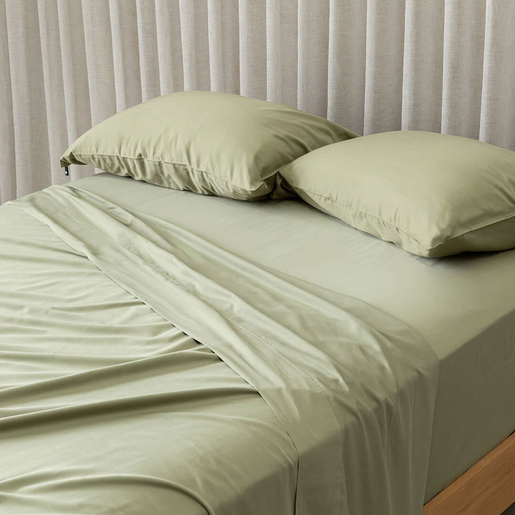 Green bed sheets and pillows on a bed with a neutral curtain background.