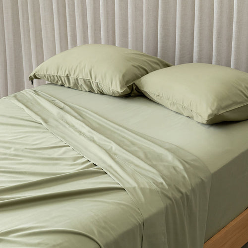 Green bed sheets and pillows on a bed with a neutral curtain background.