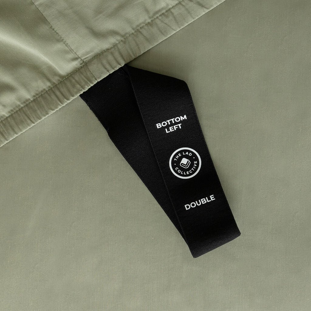 Close up of labelled corner loop strap on sage green fitted sheet that reads 'Bottom Left' and 'Double'.