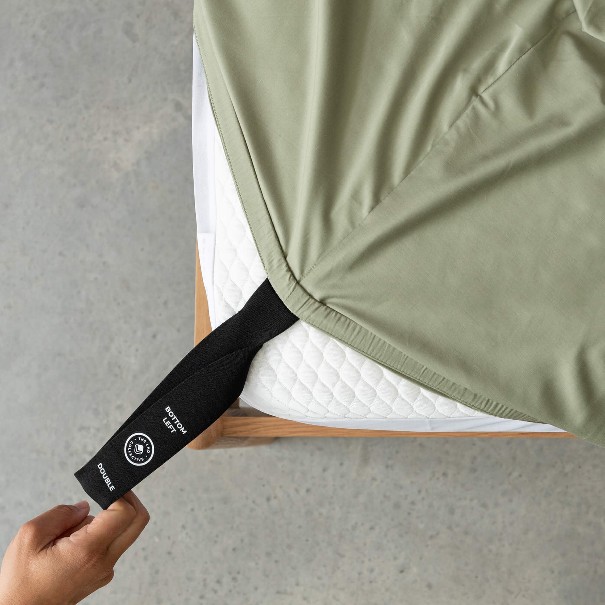Sage green fitted sheet labelled corner loop strap being pulled by a hand that says 'Bottom Left' and 'Double'.