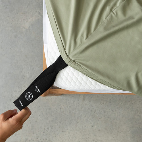 Close up of a hand pulling the labelled corner loop strap on sage green fitted sheet onto the mattress.