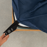 Hand pulling a black elastic corner band labeled 'Bottom Left' while securing a navy blue fitted sheet onto a mattress corner