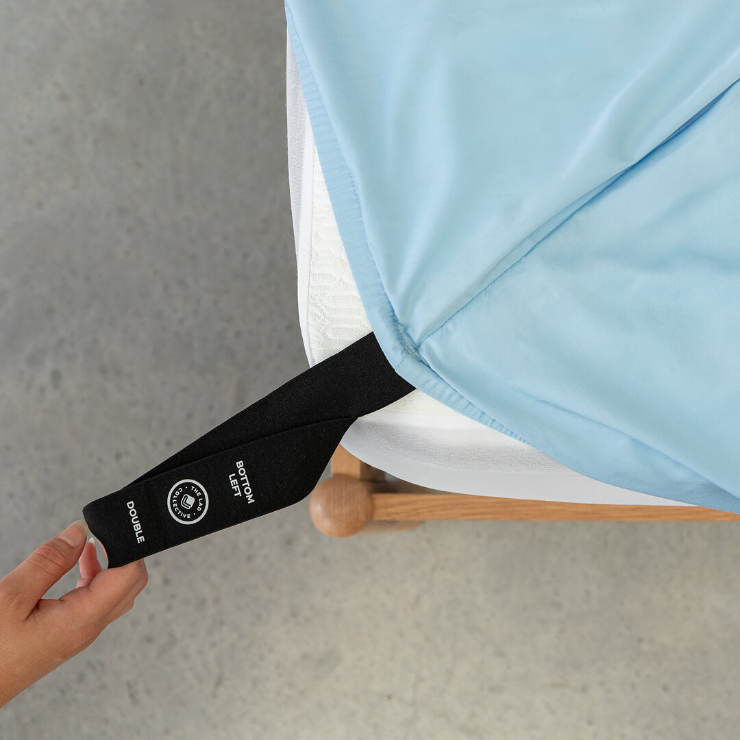 Pastel blue fitted sheet labelled corner loop strap being pulled onto bed.