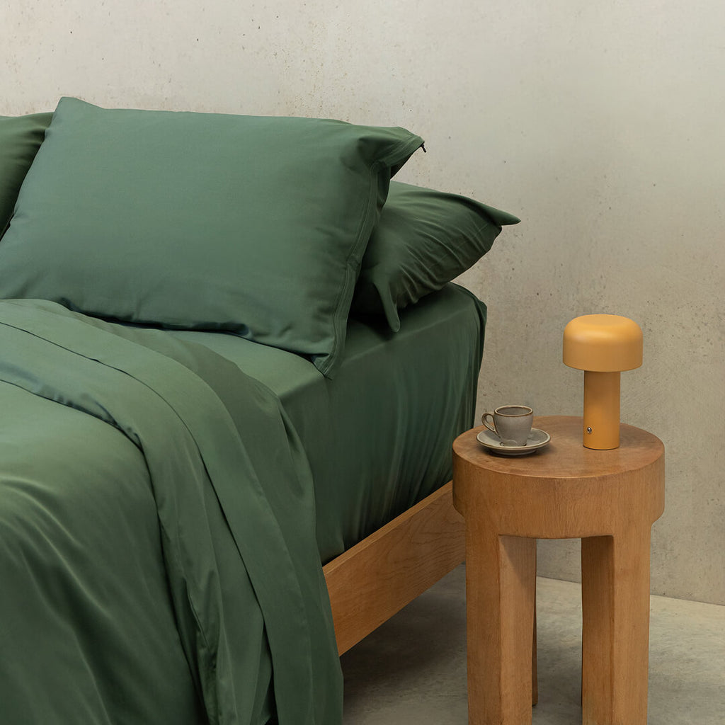 Angled shot of a dark green quilt cover on a bed, with a bedside table to the right and an orange lamp.