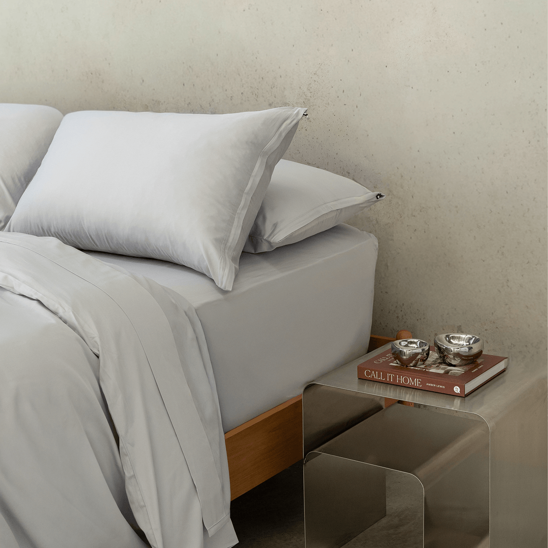Close-up of layered gray pillows and bedding beside a metallic bedside table with stacked books and decorative silver bowls.