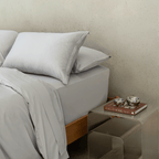 Close-up of layered gray pillows and bedding beside a metallic bedside table with stacked books and decorative silver bowls.
