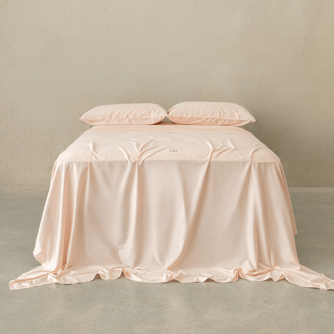 Soft blush pink fitted sheet set styled on a neatly made bed with matching pillowcases, featuring a smooth drape, elegant finish, and calming bedroom aesthetic.
