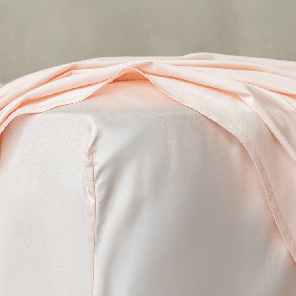 Close-up of blush pink fitted sheet fabric draped over a mattress corner, highlighting the soft texture, smooth finish, and quality construction.