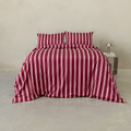 Front view of a bed styled with a burgundy quilt cover and matching pillowcases featuring vertical pale pink stripes, beside a metallic bedside table and lamp.