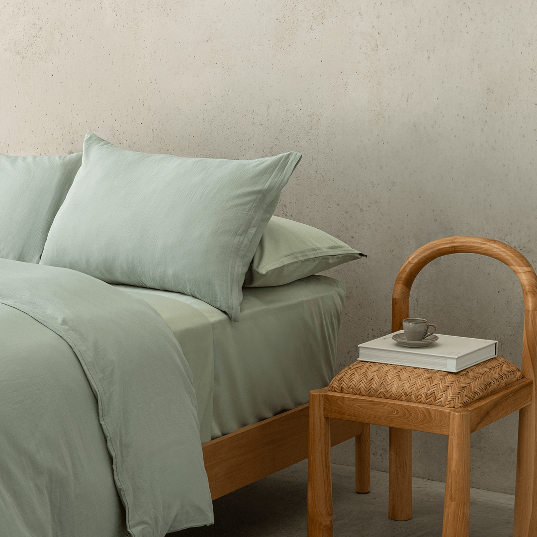 Close-up of the sage green quilt cover and pillowcases styled beside a natural timber chair, highlighting the soft washed texture and relaxed finish.