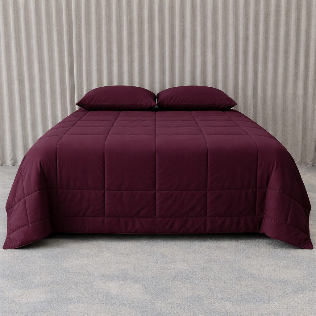 Burgundy quilted cooling blanket on a bed with pillows against a neutral background.