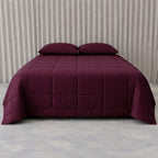 Burgundy quilted cooling blanket on a bed with pillows against a neutral background.