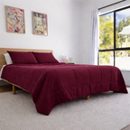 Bed with burgundy comforter and pillows in a bright bedroom with large windows.