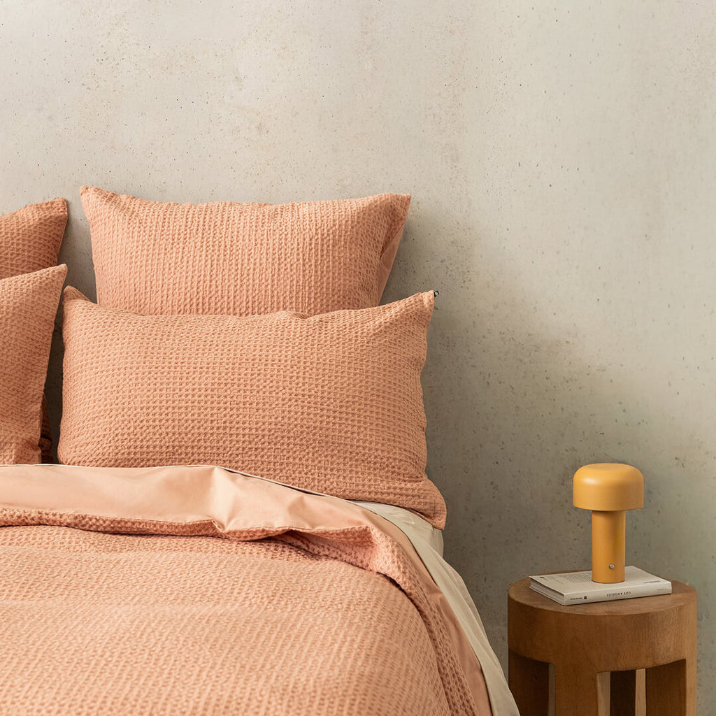 Bed with clay coloured, waffle textured bedding and pillows against a beige wall, next to a wooden side table with a lamp.