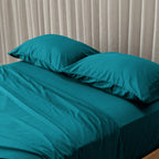 Teal bedspread and pillows on a bed with beige curtains in the background