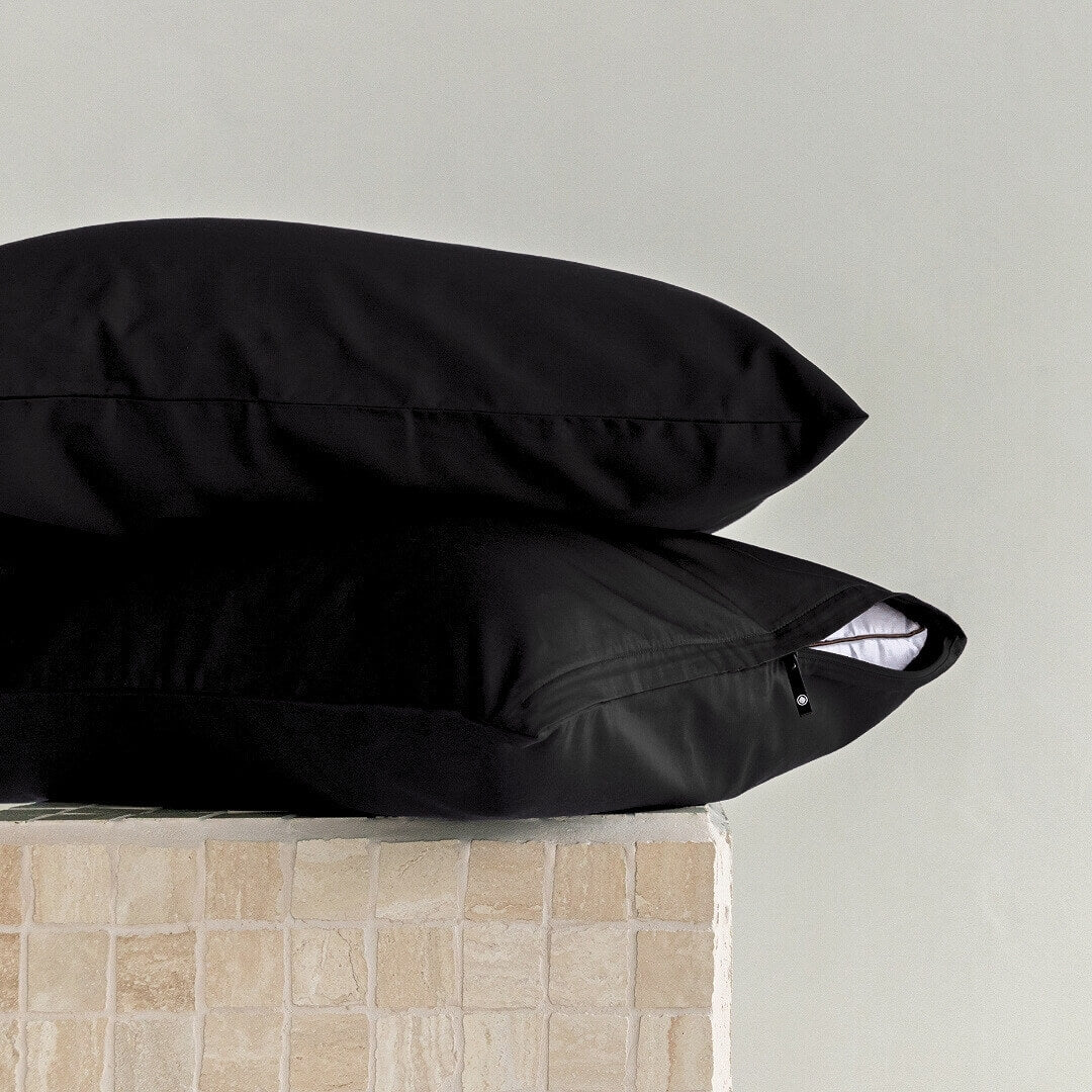 Two black pillows stacked on a light gray surface with a neutral-colored wall in the background.