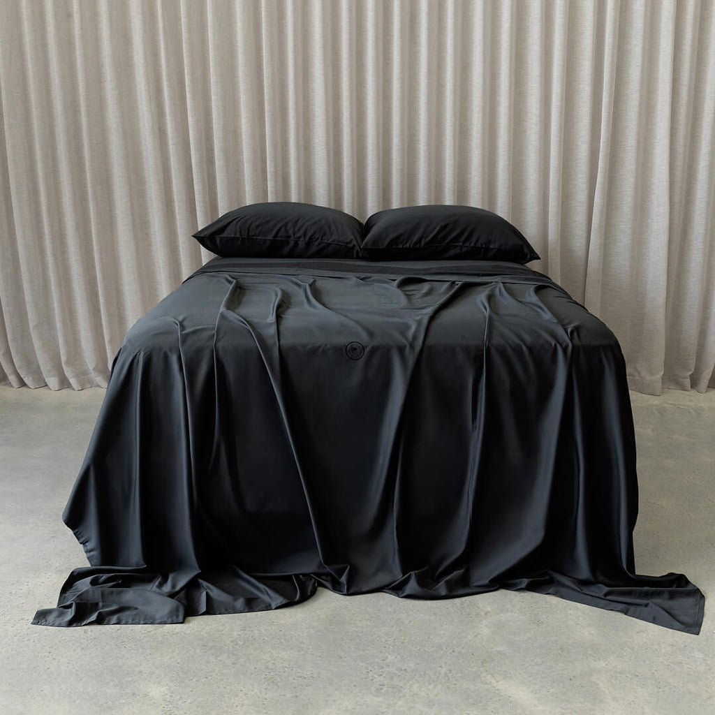 Black bedspread on a bed with pillows against a beige curtain background.