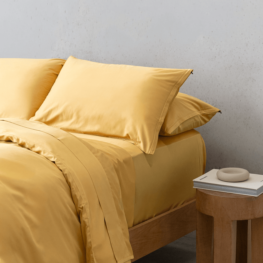 Side view of a double bed styled with a golden yellow quilt cover, matching pillowcases, and a wooden nightstand