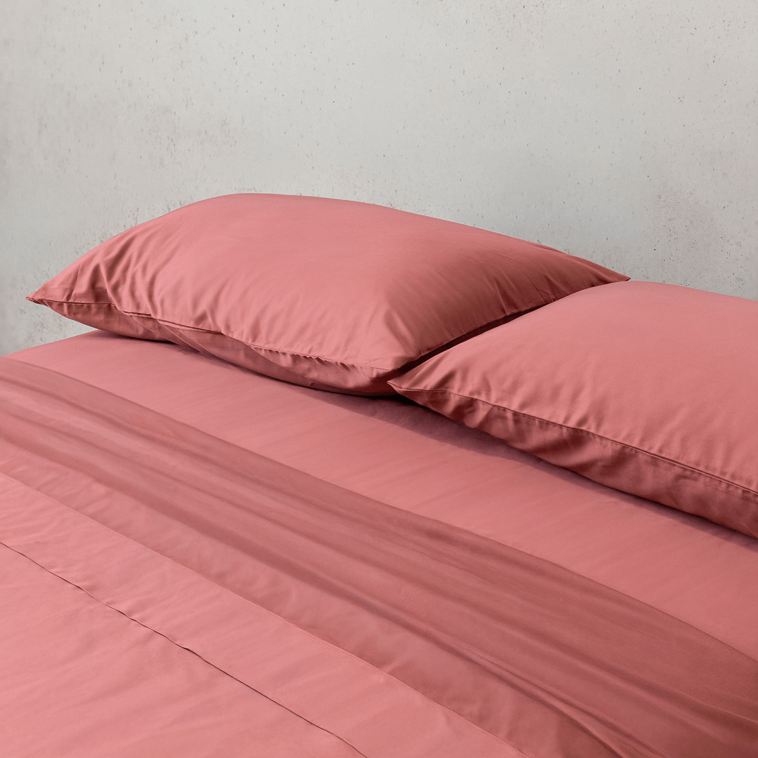 Angle view of two dusty rose pink pillowcases and a matching flat sheet neatly arranged on a bed