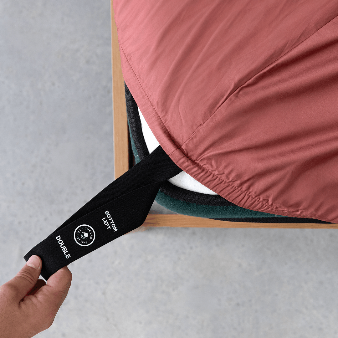 Hand pulling a black elastic corner band labeled 'Bottom Left' while securing a dusty rose pink fitted sheet onto a mattress corner
