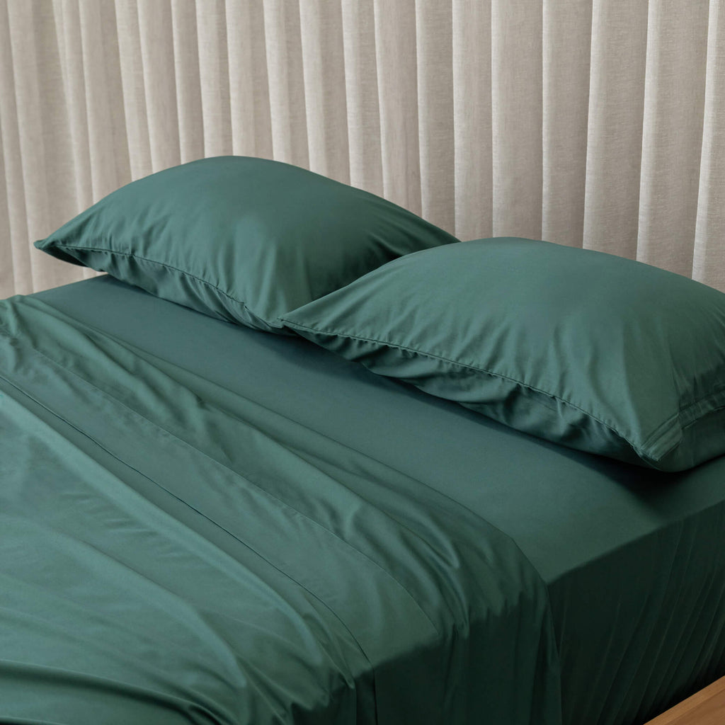 Green bed sheets and pillows on a bed with beige curtains in the background