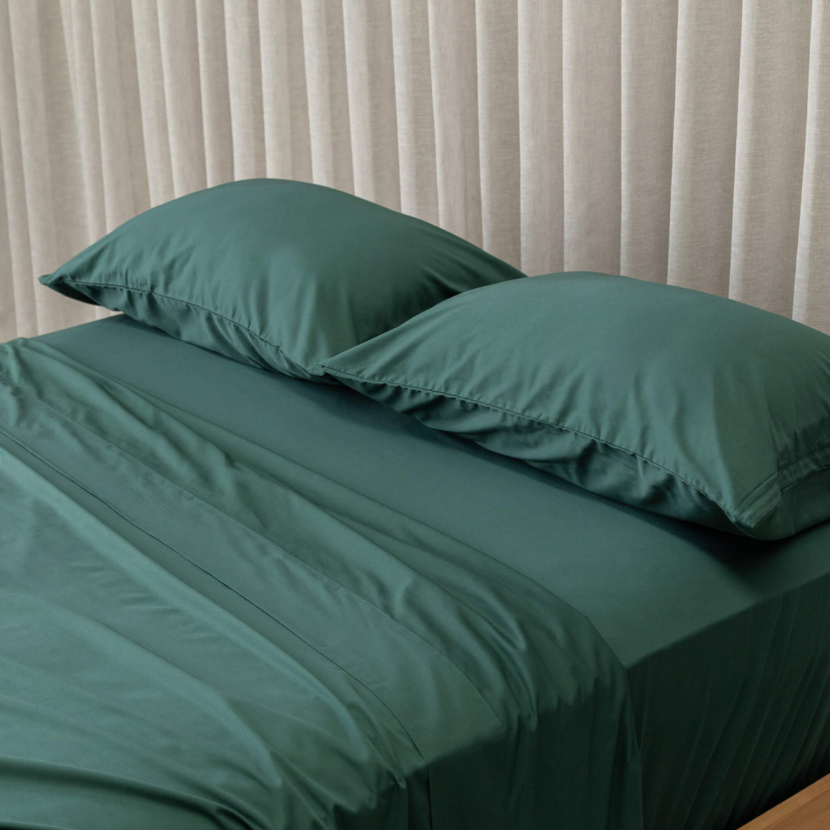 Green bed sheets and pillows on a bed with beige curtains in the background