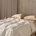Beige quilt insert and pillows on a bed with a neutral wall and curtain in the background.