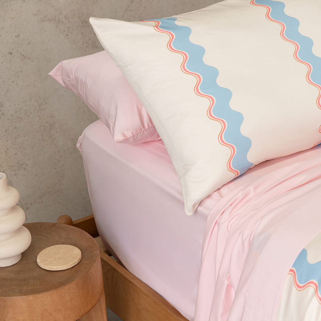 Bed with pink bedding and a decorative pillow with wavy pattern, wooden side table with white vase and coaster.