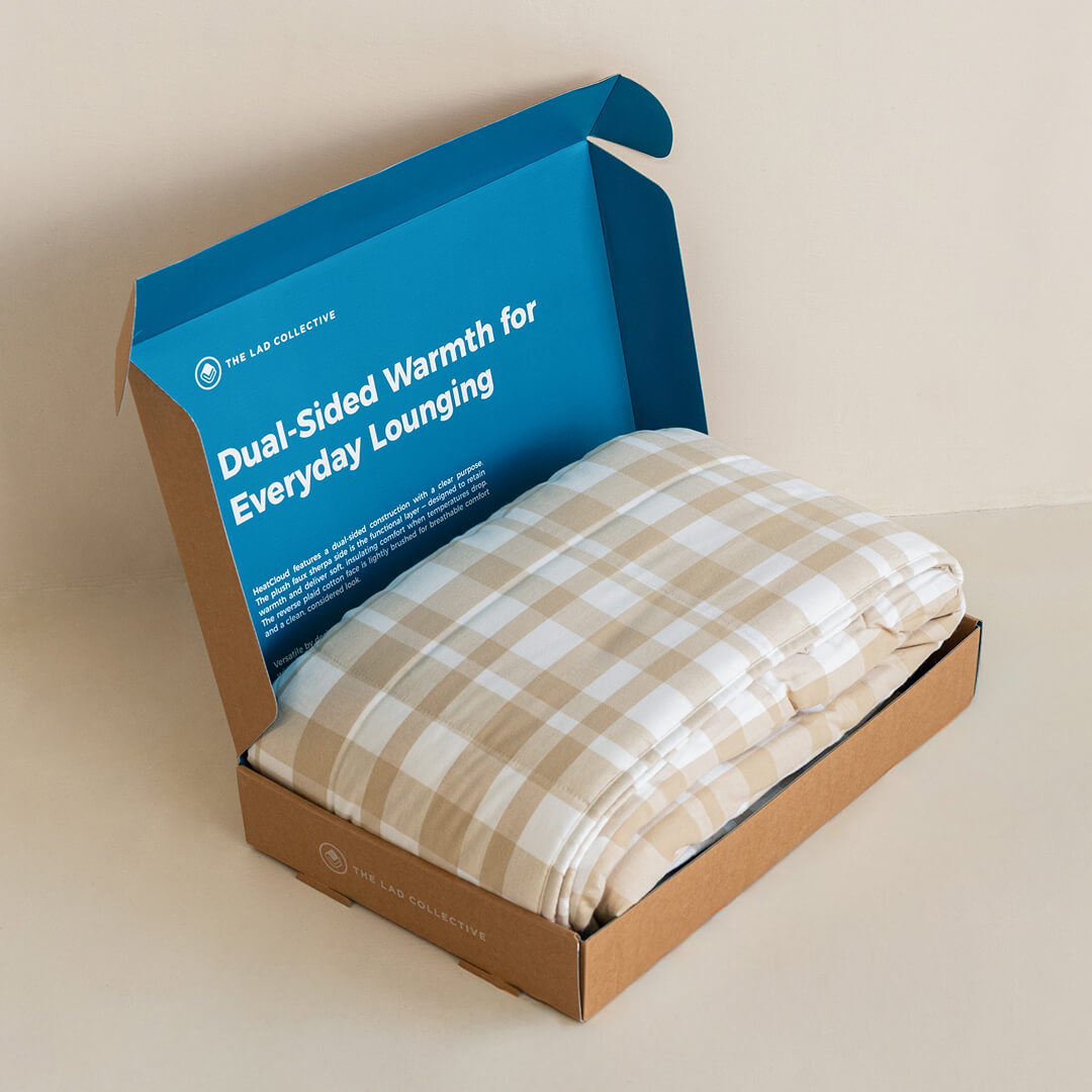 Folded checkered blanket in an open box with blue lid displaying text on a beige background.