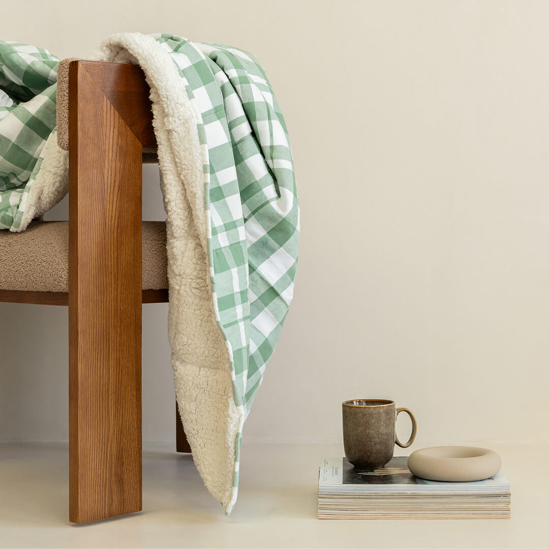 Green checkered blanket draped over a wooden chair with a cup and book on a table.