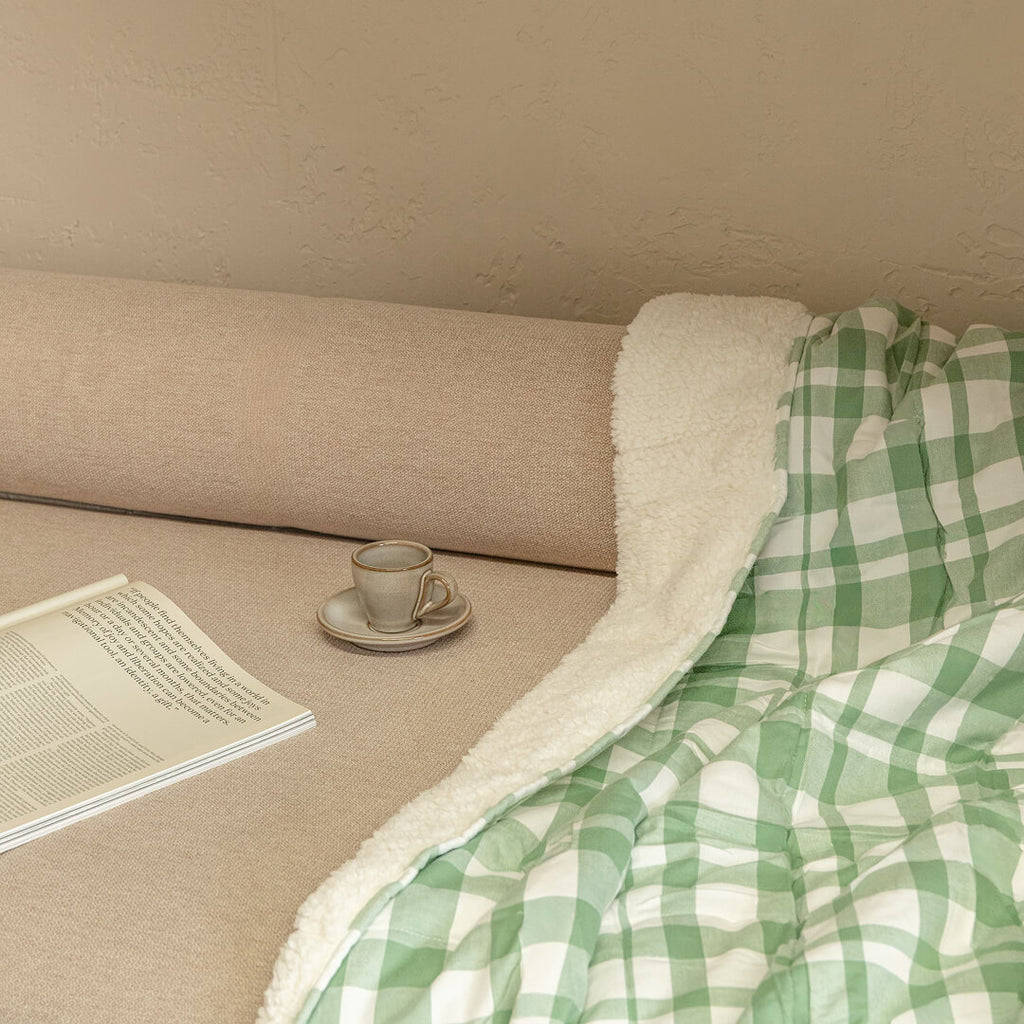 Green and white checkered blanket draped over a beige couch with a cup and saucer, and a book.