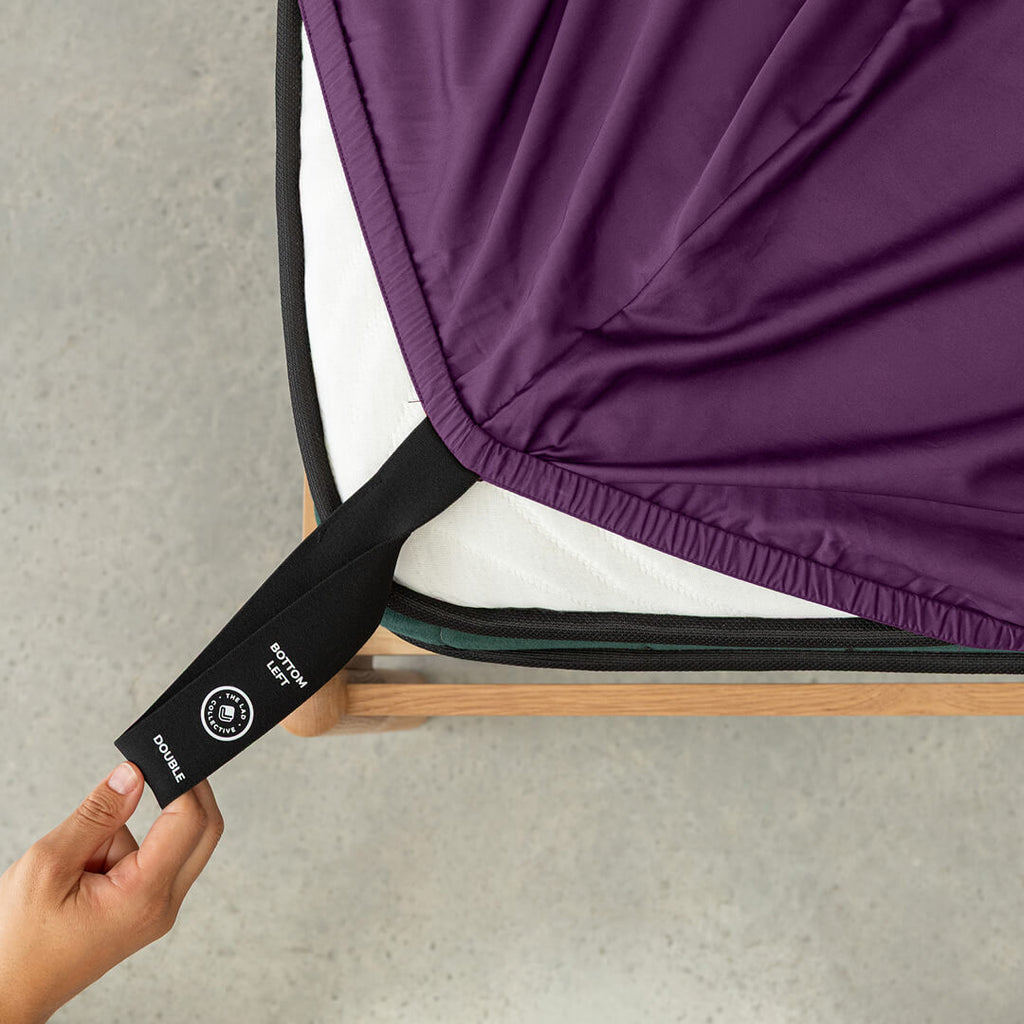 Deep purple fitted sheet being pulled onto mattress using labelled corner loop strap.