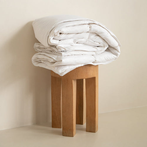 Folded white summer quilt insert on a wooden stool against a beige wall.