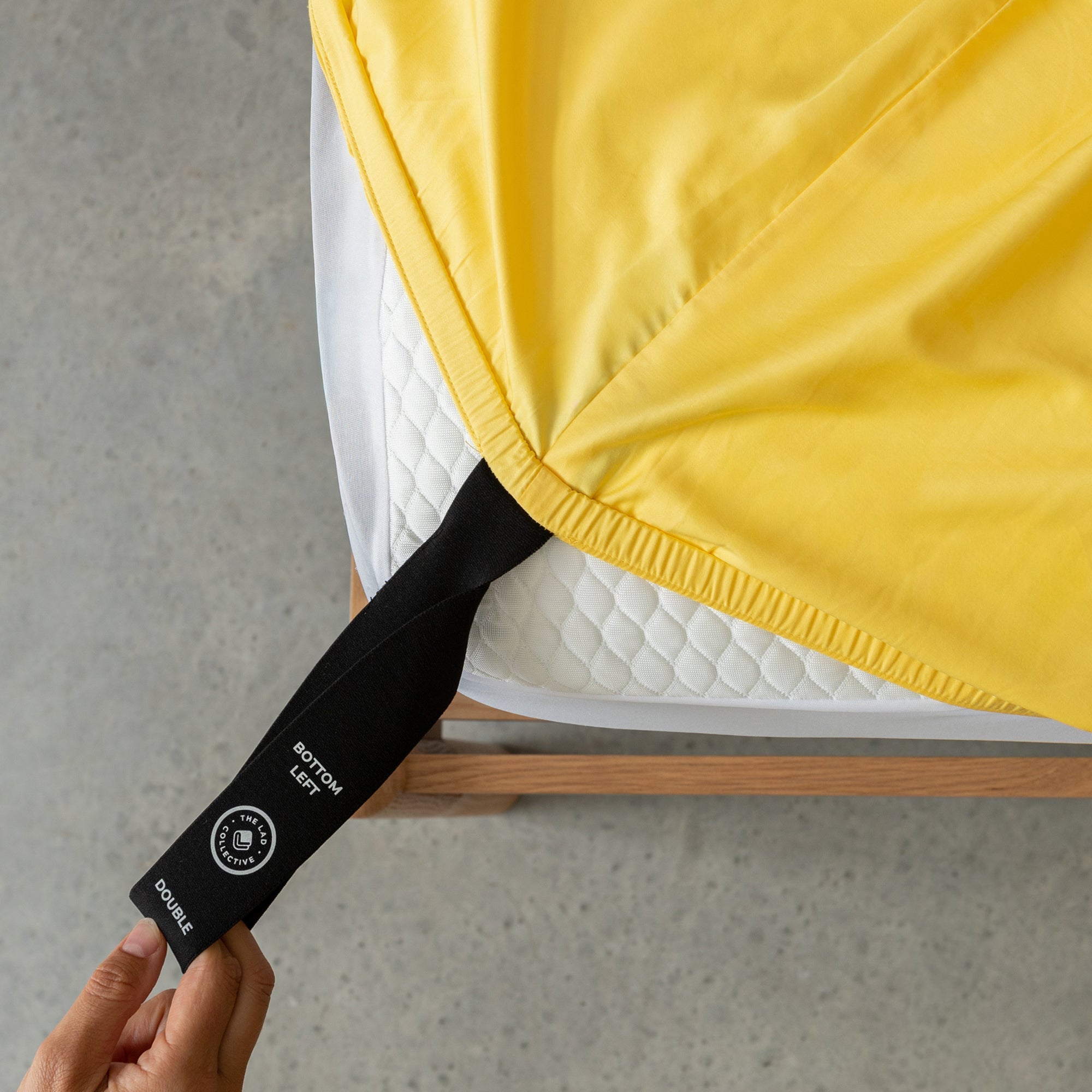 Close-up of a hand pulling the labelled corner loop strap of a bright yellow fitted sheet over the corner of the mattress. 